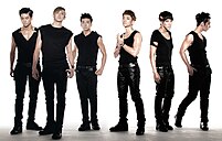Six men wearing form-fitting black sleeveless shirts, leather pants, and combat boots. They have prominent eye makeup and each has a different gelled hairstyle.