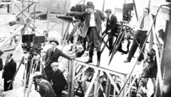 DeMille on a stand, holding a megaphone, giving commands to crew members which surround him and several film cameras