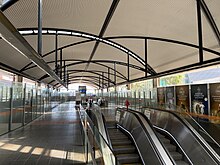 Tiled concourse with glass walls and a curved roof. There are escalators and a lift for transport to platform level.