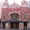 A red brick building with buff terracota dressings; Victorian Baroque in style. Two storeys with a three bay front. Large, illuminating wording of "Hackney Empire" on the front, in red lettering.