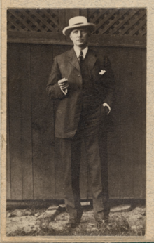 A tan and black image of a man posing in front of a barn with a white hat and suit