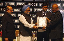 Four men in formal attire are standing on a stage in front of a black background with white text on it. The two men in the middle are both grabbing an award plaque.