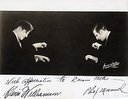 Two men opposite, hands raised above invisible keyboards, signatures below in bottom right; another unidentified signature in the bottom right of the picture.