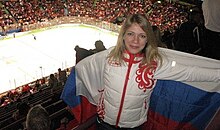 A woman with shoulder-length blonde hair is wearing a white jacket with a red pattern. She is hold a Russian flag behind her back with white, blue and red horizontal stripes. Below her is a horizontal ice rink and a large group of seats is surrounding it from all sides, some with spectators occupying them.