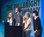 Four smiling people (two women and two men) stand in front of a podium. Behind them is a large blue screen with the words THE FULLBRIGHT COMPANY projected across it.