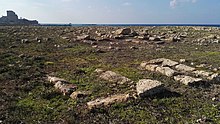 Pierres éparpillées dans un champ d'assez grande surface, situé à proximité des ruines d'un château, au bord de la mer.