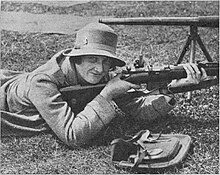 Badcock, lying in the prone position with a Lee-Enfield rifle, on a grass range. She wears tweed clothing, including a hat and jacket, and has a small satchel open beside her.