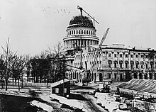 A black and white photograph of the United States Capitol building, showing construction equipment and an unfinished dome