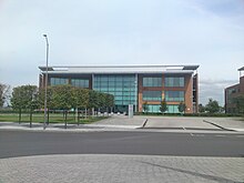 A wide orange rectangular building with turquoise windows, partly obscured on the left by rows of trees and a road light