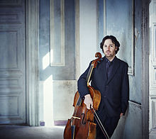 photo : un homme en costume, tenant son archet et portant son violoncelle sous le bras.