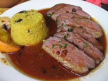 Photographie couleur montrant une assiette blanche garnie. Sept tranches de magret de canard cuites saignantes, baignent dans un jus de viande rouge avec une timbale de semoule démoulée et un quartier d'orange. Le tout est saupoudré de persil.