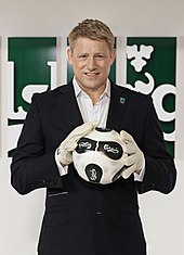 Blonde haired white man in a black suit with white gloves holding a black and white football.
