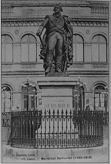 Photographie ancienne d'une statue en pied d'un maréchal de Napoléon en grand uniforme, tenant son sabre de la main gauche et son bâton de maréchal de la main droite. Autour du piédestal, une grille, et en arrière-plan, un bâtiment.