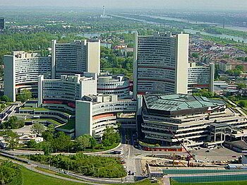 Huge complex of skyscrapers and other large buildings interlaced with trees and gardens. The surrounding area as far as the horizon is filled with trees and large rivers. In the foreground a crane and small building site show that a new construction is underway.