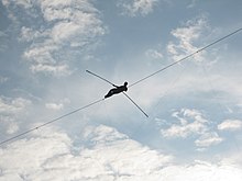 A small figure holds a balancing pole while lying on a high-wire