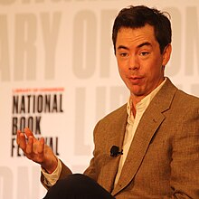 A photograph of Kyle Chayka. He is a white man with a slight widow's peak, high eyebrows and pursed lips. He is waring an open-collared white shirt and a brow plaid suite jacket. He is in front of backdrop printed with the Library of Congress National Book Festival logo.