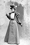 Black-and-white photograph of a young black woman wearing a hat with feathers and a suit: a coat with with long, leg-of-mutton sleeves and wide-black lapels with a skirt the same cloth as the coat. The coat's skirt passes just over her hips. The coat is open to show a white blouse beneath. The woman is holding a cane behind her back.