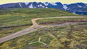 An aerial view of the emergency backcountry helicopter landing zone located at the summit of Rollins Pass; the white prong of the landing zone, furthest from the camera, points north. The comparatively low saddle of Rollins Pass is visible in this image as the summit itself (in the foreground and midground) is plainly lower in elevation than the surrounding mountains. The wood debris consists of both remnants from snowsheds that covered the tracks as well as discarded railroad ties that were removed from service in the summer of 1936.