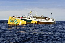 Yellow and white wave energy device in a calm sea. It is an asymmetric boat hull, leaning towards the photographer. The name Wello and a penguin are painted on the site.