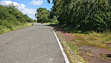 Narrow single carriageway, with wide grass verges, in a flat rural landscape