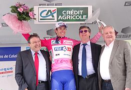 Quatre hommes sont alignés. Le deuxième, vêtu d'un maillot rose, lève les bras avec un trophée et un bouquet.