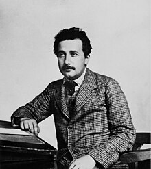 Head and shoulders shot of a young, moustached man with dark, curly hair wearing a plaid suit and vest, striped shirt, and a dark tie.