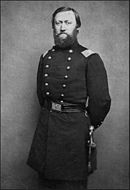 Black and white photo shows a bearded man standing. He wears a dark military uniform with two rows of buttons.