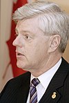 A headshot of John Manley from the side, in a suit, backgrounded by a flag