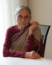 A photograph of an old woman sitting on a chair wearing sari with right hand kept on the table