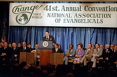 Reagan, smiling, standing behind a lectern with the presidential seal and a microphone. Behind him is a banner for the convention, with the slogan "Change Your World" and the title "41st. Annual Convention NATIONAL ASSOCIATION OF EVANGELICALS". Behind the banner are curtains. In front of the banner and curtains but behind Reagan are two rows of seated people, about 20 total. Most seem in good spirits with smiles and expressiveness. Most are Caucasian men. Two women are visible, as is a Black man.