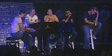 Backstreet Boys sitting on stools with microphones and singing, with one of them playing the piano.