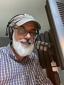 Radio host John Dankosky, in front of a studio microphone, hosting Science Friday, while wearing a Hartford Whalers hat.