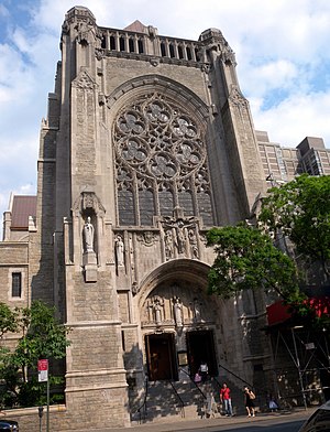 A brown stone tower with rosette window and other decorative touches rising up from a street with trees