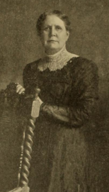 An older white woman, standing with her hands on the back of a carved wooden chair; she is wearing a dark garment with a high white lace collar