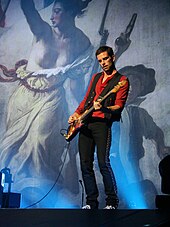 A short-haired man wearing a red shirt and a black waistcoat plays the bass as a picture of Eugène Delacroix's Liberty Guiding the People appears in the background