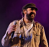 A man with a black hat and a light brown jacket is holding a microphone on his right hand