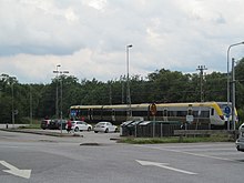 A small parking lot with many streetlights seen diagonally from across an intersection, with traffic signs in front of it, one of which is a red circle containing the number "50" on a yellow background. Behind the parking lot is a short modern-style gray, yellow and blue streamlined train underneath overhead wires.