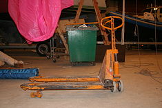 Jack showing how wheels drop, lifting forks and load. The steering tiller and raised hydraulic cylinder are clearly visible on the right