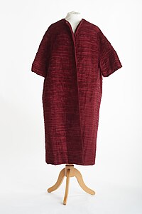 Color photograph of a burgundy-colored, very loose fitting coat that opens down the middle, reaches the wearer's knees and has short loose-sleeves that stop before the elbow. There is a rough appearance to the texture of the cloth.