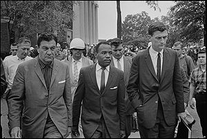 Chief U.S. Marshal James McShane and U.S. Assistant Attorney General for Civil Rights John Doar are pictured escorting James Meredith to class at Ole Miss after the riot. Large groups of federal agents and likely students are seen in the background.