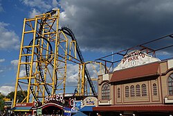 View of the Steel Curtain roller coaster from a nearby path. The roller coaster contains yellow supports and black track. A building is to the right.