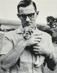 A man looking down at a lion cub, who he holds carefully against his chest.