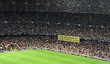 A section of the stands in a football stadium, with a capacity crowd. A corner of the pitch is visible at bottom. A large yellow banner hangs from the second tier of the stands, which says "Only dictatorships jail peaceful political leaders" (in English) in block capitals.