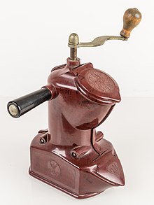 un moulin à café en bakelite, de couleur brun clair, avec une poignée latérale en bakélite grenat, et avec une manivelle en métal pour actionner un axe métallique vertical