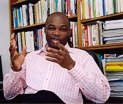 Charles Onana wearing black pants and a pink-and-white checkered shirt, sitting in a chair with bookcases in the background, appearing to speak and gesticulate while looking at camera