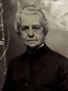 An older white woman with white hair in an updo, wearing an all-black top with a high collar