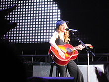 Image of a blonde woman sitting on a stool. She's wearing a black hat, black sleeveless shirt and black pants. She's holding an orange acoustic guitar in her hands. A microphone stands before her. Gray bright imagery can be seen in the background.