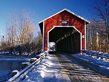 Une photographie en couleurs représentant, dans un paysage enneigé, un pont couvert vu dans l'axe de celui-ci. Son porche est de couleur rouge vif cerclée de blanc.