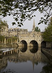 A three arch stone bridge with buildings on it, over water. Below the bridge is a three step weir and pleasure boat.