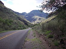 RM 1832, a two-lane road, winds through a canyon with steep hillsides on both sides of the road. In the distance are rugged mountains with steep cliffs covered with desert grasses and brush.
